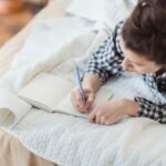 Femme écrivant dans un carnet, installée sur son lit