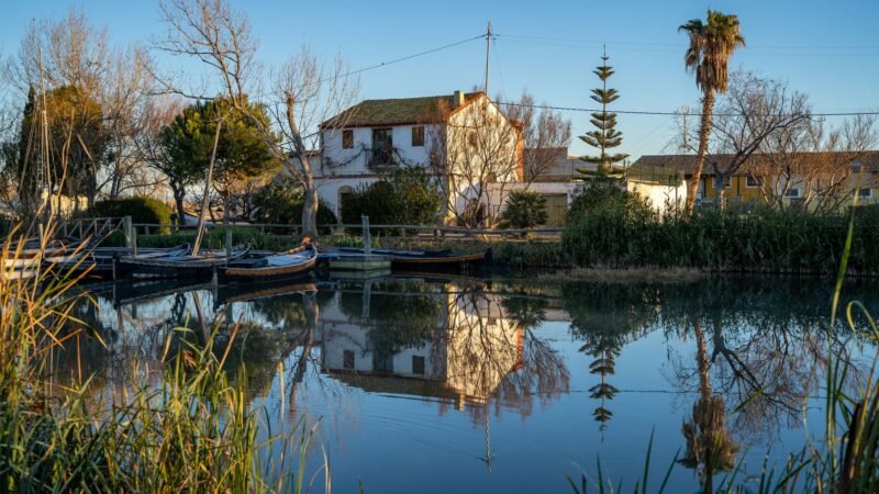 Une maison blanche et des barques amarrées se reflètent dans l'eau calme d'un canal de l'Albufera, près de Valence